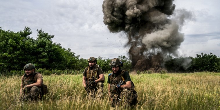 Ukrainian soldiers taking cover while the smoke of the explosives detonated rises in the background