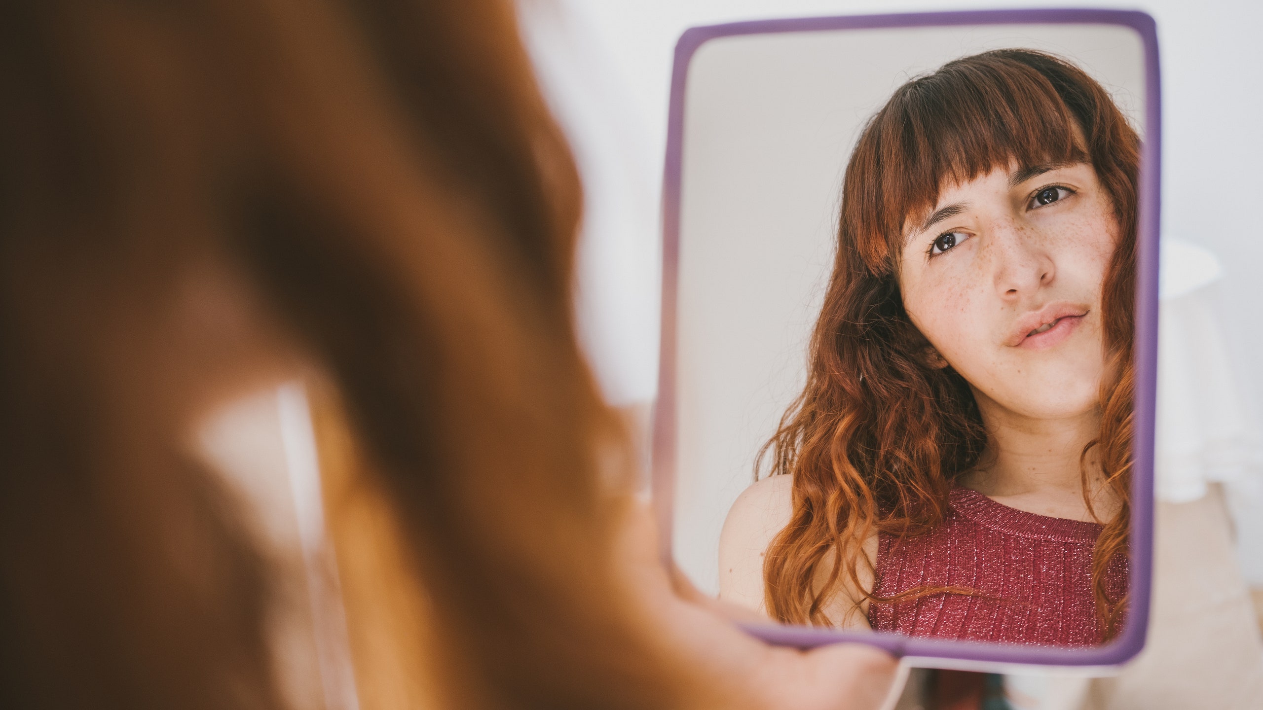 person with bangs and curly red hair looking into a handheld mirror