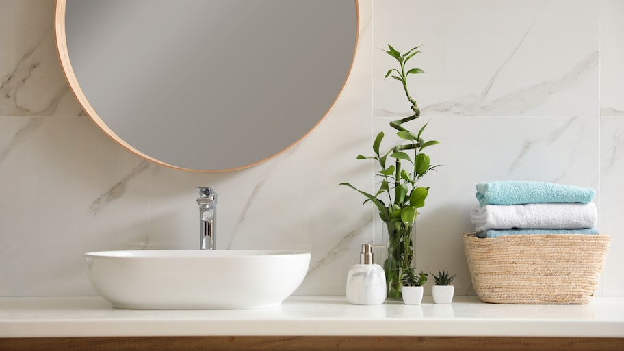 Modern bathroom with marble slab backsplash, round mirror, plants, vessel sink, and a woven basket with towels