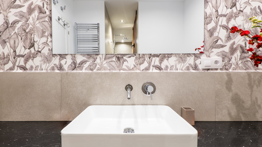 Modern bathroom with black countertops, white sink, chrome fixtures, square mirror, and wallpaper backsplash