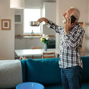 text:Angry Bearded Senior Man Talking on Smartphone Near Plastic Wash Bowl at Home in the Living Room Because of Roof Leaking