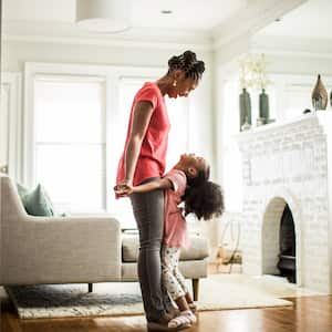 Girl standing on top of mother’s feet in contemporary living room with white fireplace and greige chair