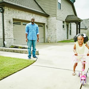 Smiling mother and father watching young daughter ride scooter in driveway of home