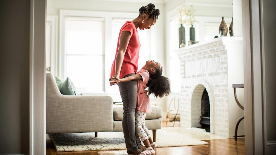 A girl standing on her mother’s feet in a living room with a fireplace