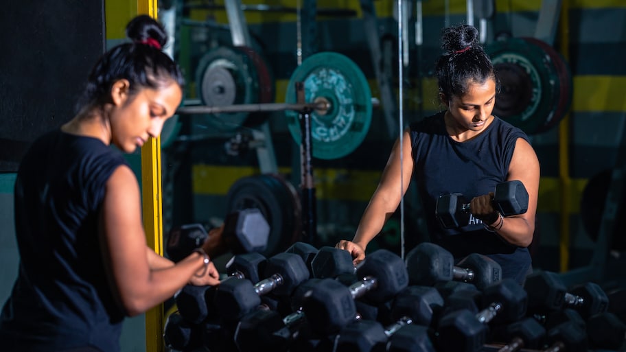 Woman leaning on weight rack with a dumbbell in her right hand doing a curl facing a large mirror in her garage home gym