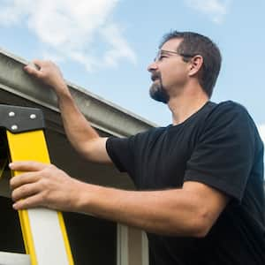 Man Inspects Roof Condition of House on a ladder