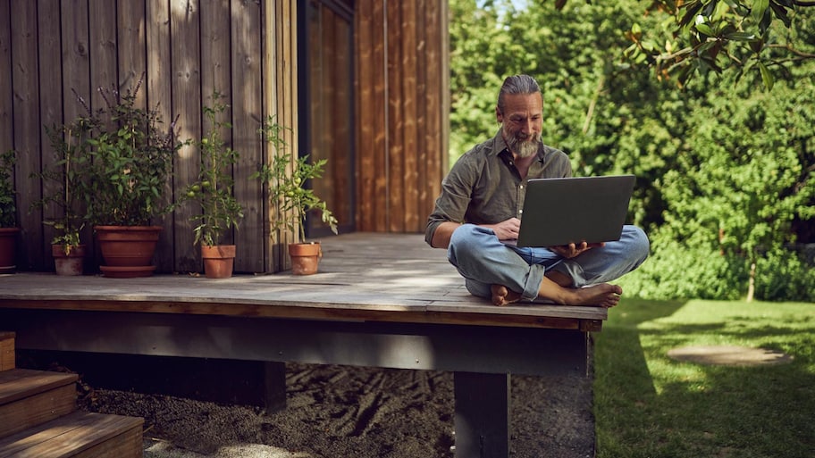 A mature man using a laptop while sitting outside a wooden house