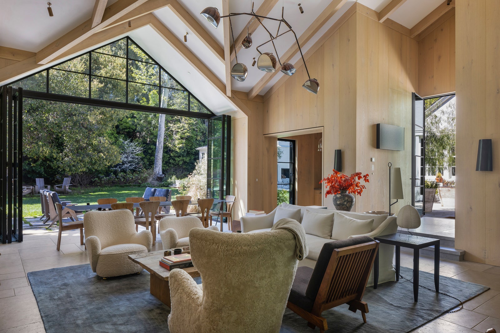 vaulted white ceiling with blonde wood beams gray rug on floor open glass wall to backyard cream sofa two beige chairs...