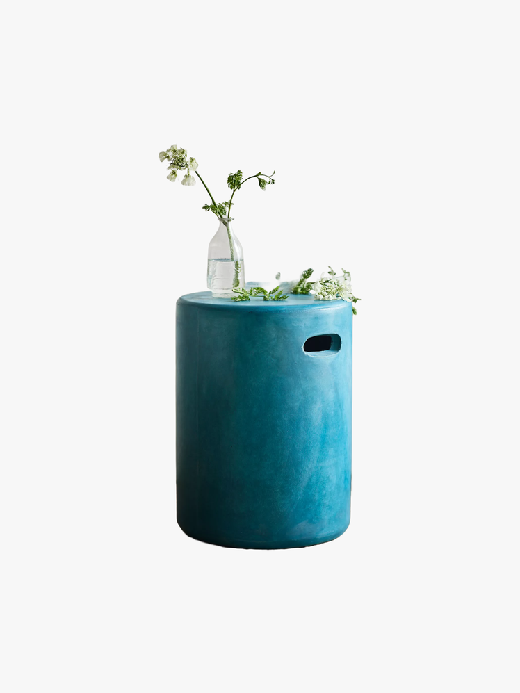 Cylindrical concrete stool with a notch carrying handle painted turquoise with plants on top of it.