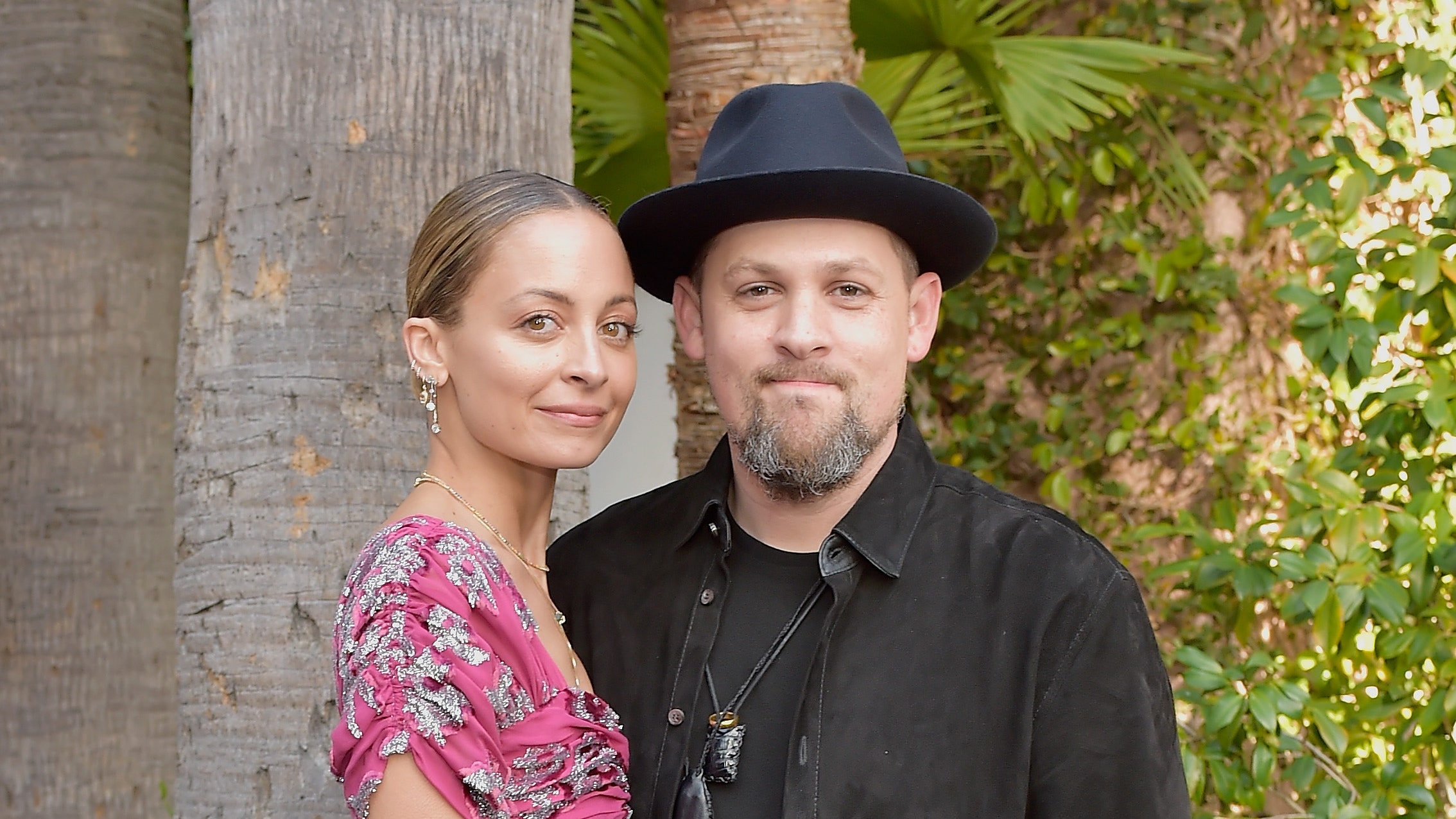 Nicole Richie left in a pink dress beside Joel Madden wearing black posed standing outside in front of trees landscaped...