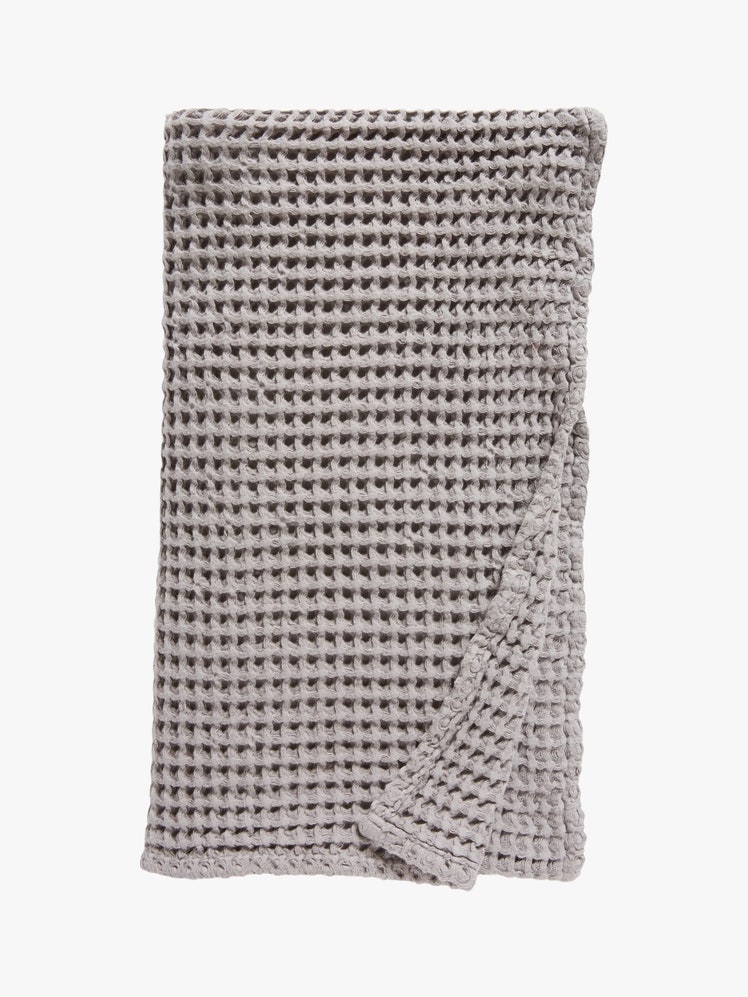 Gray bath towel with a waffled texture folded into a rectangle with the bottom right corner pulled up to reveal the...