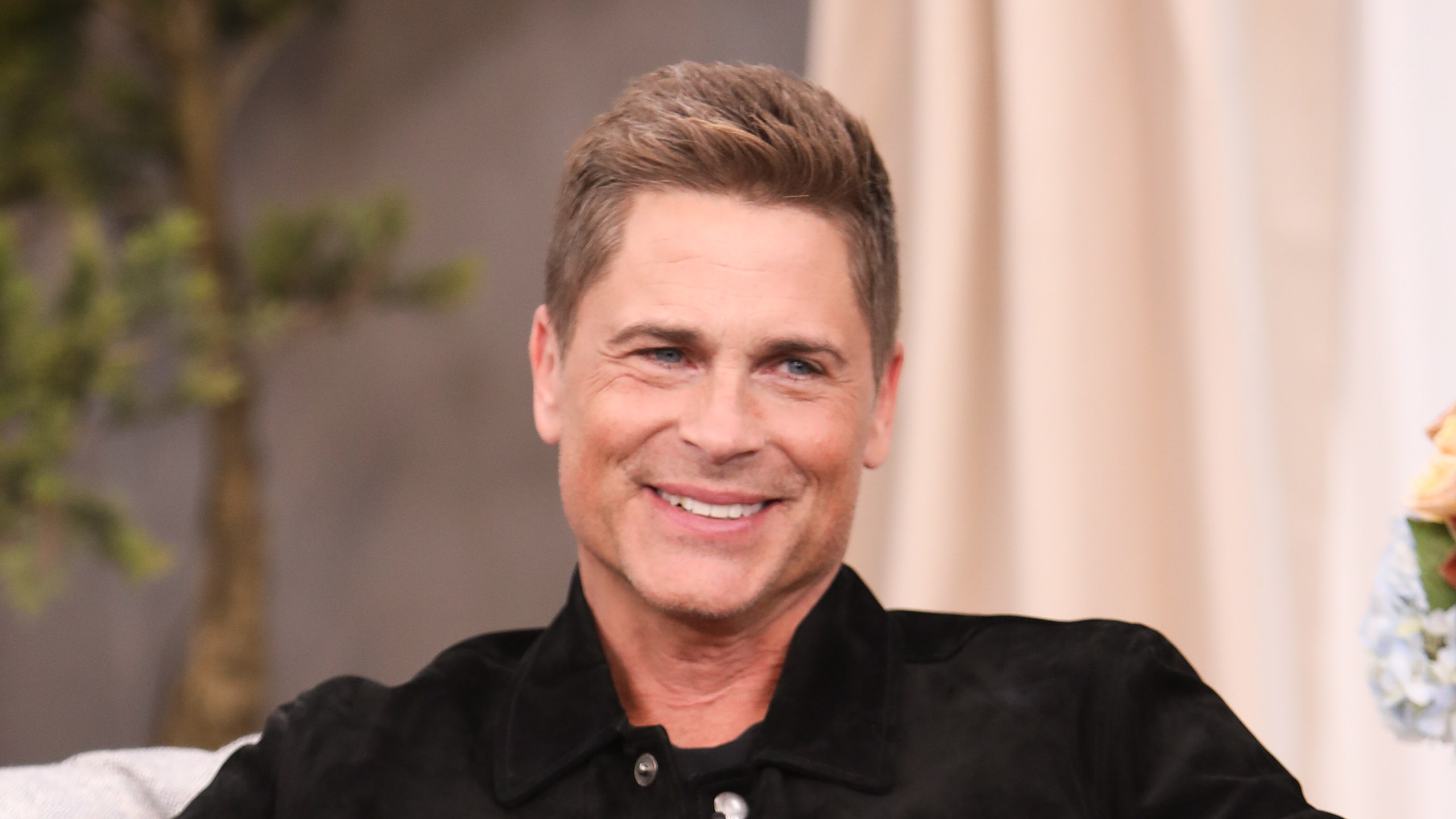 Rob Lowe in black jacket smiling seated on gray sofa