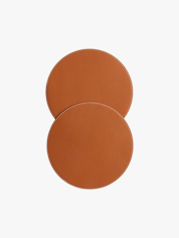 Pair of burnt orange round placemats in coated fabric with contrasting stitching at edge.