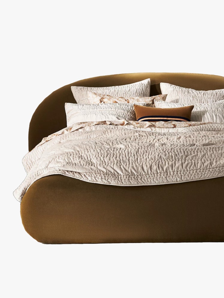 Bed with a rounded olive green bed frame made up with an offwhite quilt with stitched and textured embellishments and...