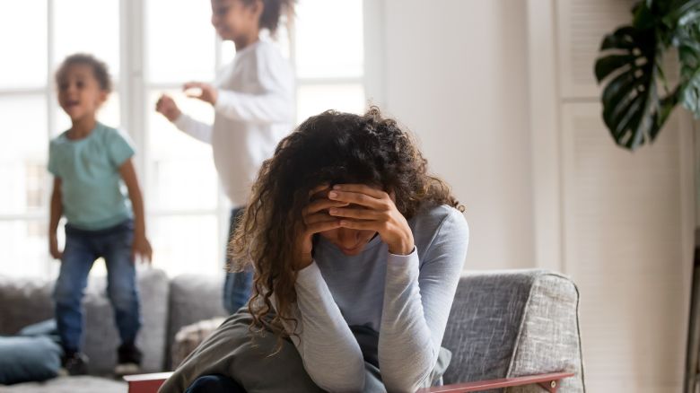 Black single frustrated woman hold her head with hands sitting on chair in living room, playful kids jumping on couch on a background. Tiredness, depression difficult to educate children alone concept