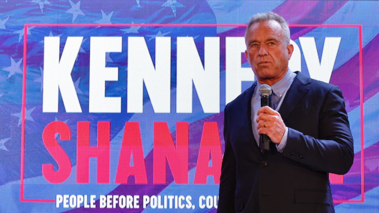 American politician Robert F Kennedy stands holding a microphone, in front of a large red, white and blue screen bearing his name. 