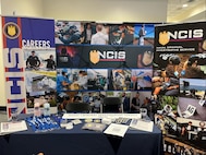 NCIS recruiting display features standing backgrounds with images of NCIS agents and personnel along with a table of giveaway items like patches, lanyards and printable materials.