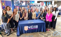 NCIS personnel pose in front of NCIS recruitment display and table at WIFLE conference.