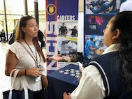 NCIS staff member speaks to attendee at the WIFLE annual conference in front of NCIS recruiting display and table.