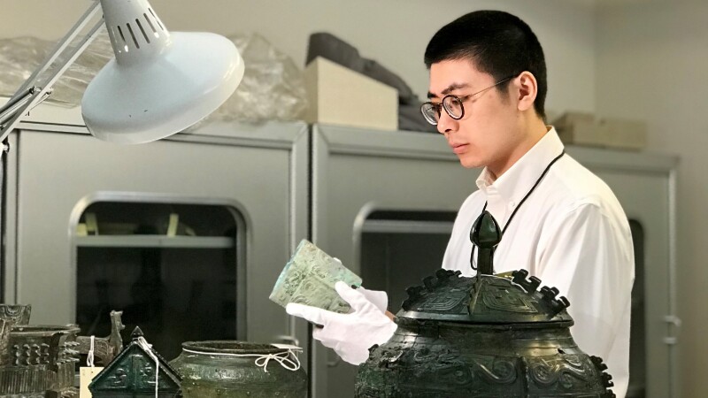 Person wearing glasses, white shirt, and gloves observing Chinese bronzes on a table.