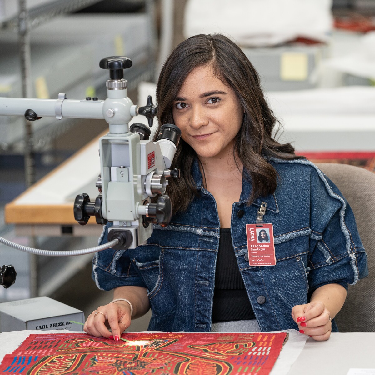 Alejandra sits at a table with a flat red art object on it. An instrument on a long arm hovers above it, casting a light on it.