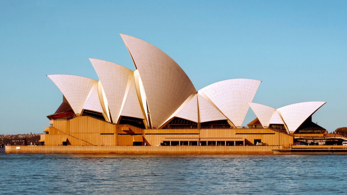 A large building with a distinctive roof shaped like ship sails sits alongside a large body of water.