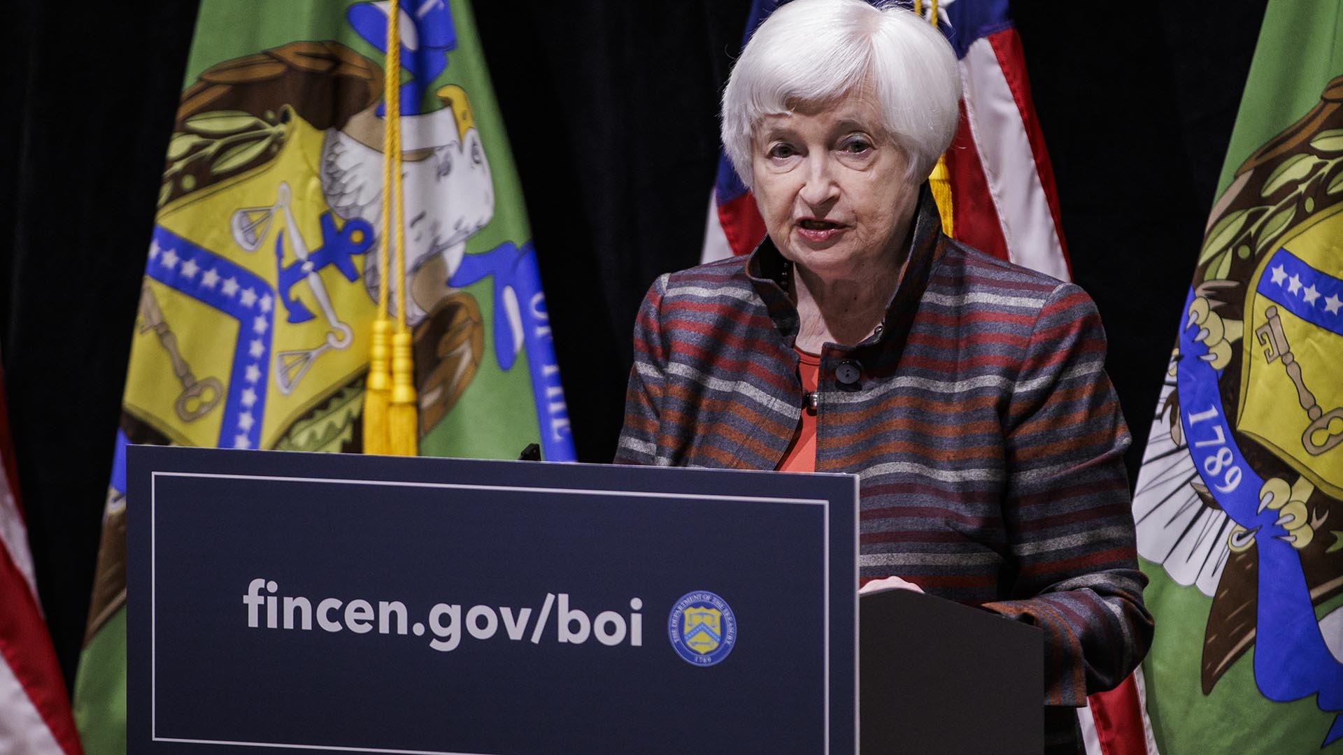 Woman stands at a podium that reads fincen.gov/boi, in front of U.S. and U.S. Treasury flags.