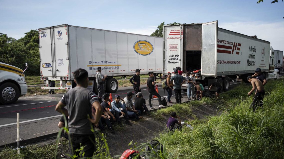 A truck trailer with an open door on the side of a road, with a row of people sitting on the ground behind the truck.