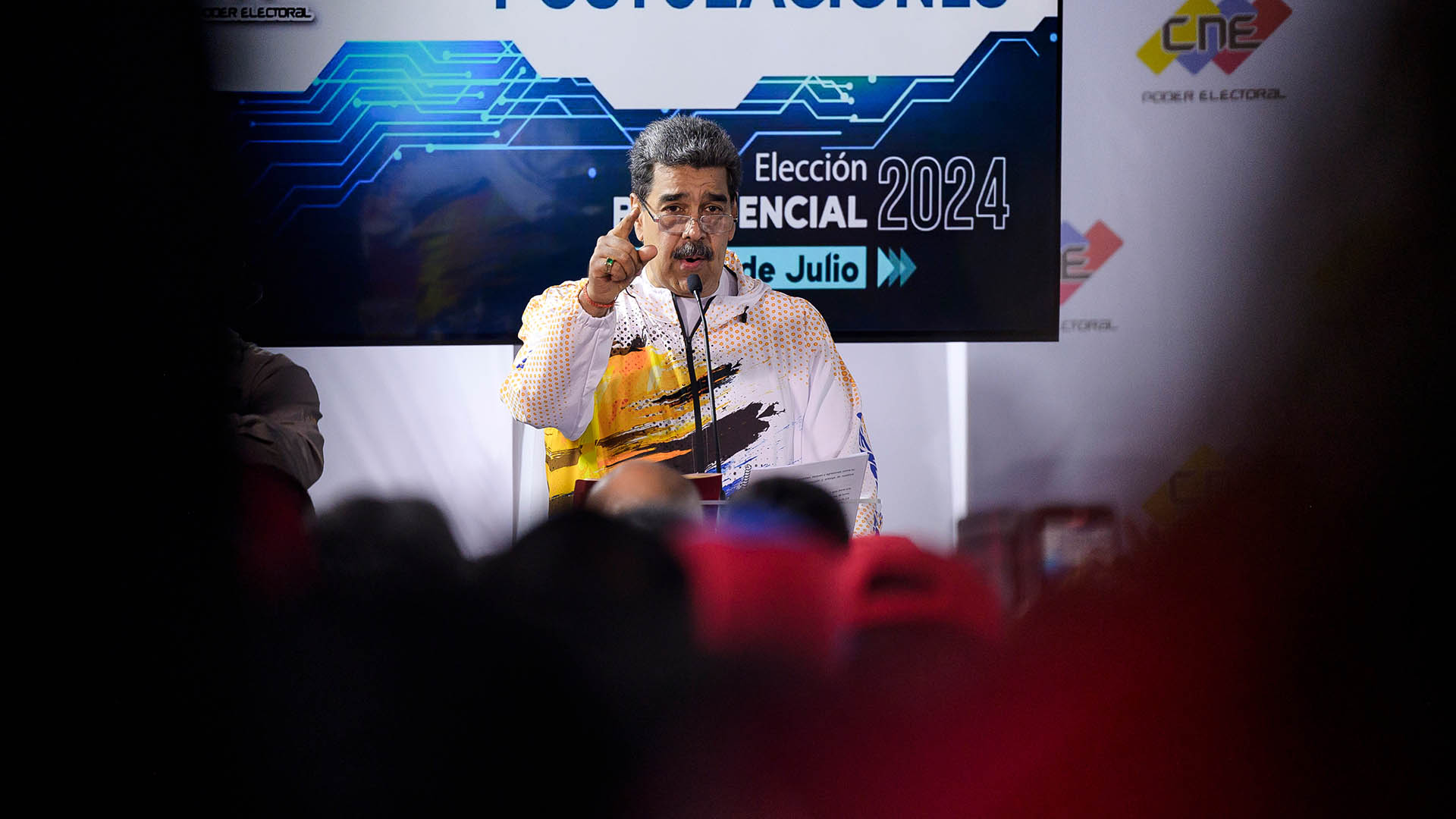 Nicolas Maduro before a silhouetted crowd, with a 2024 election sign behind him.