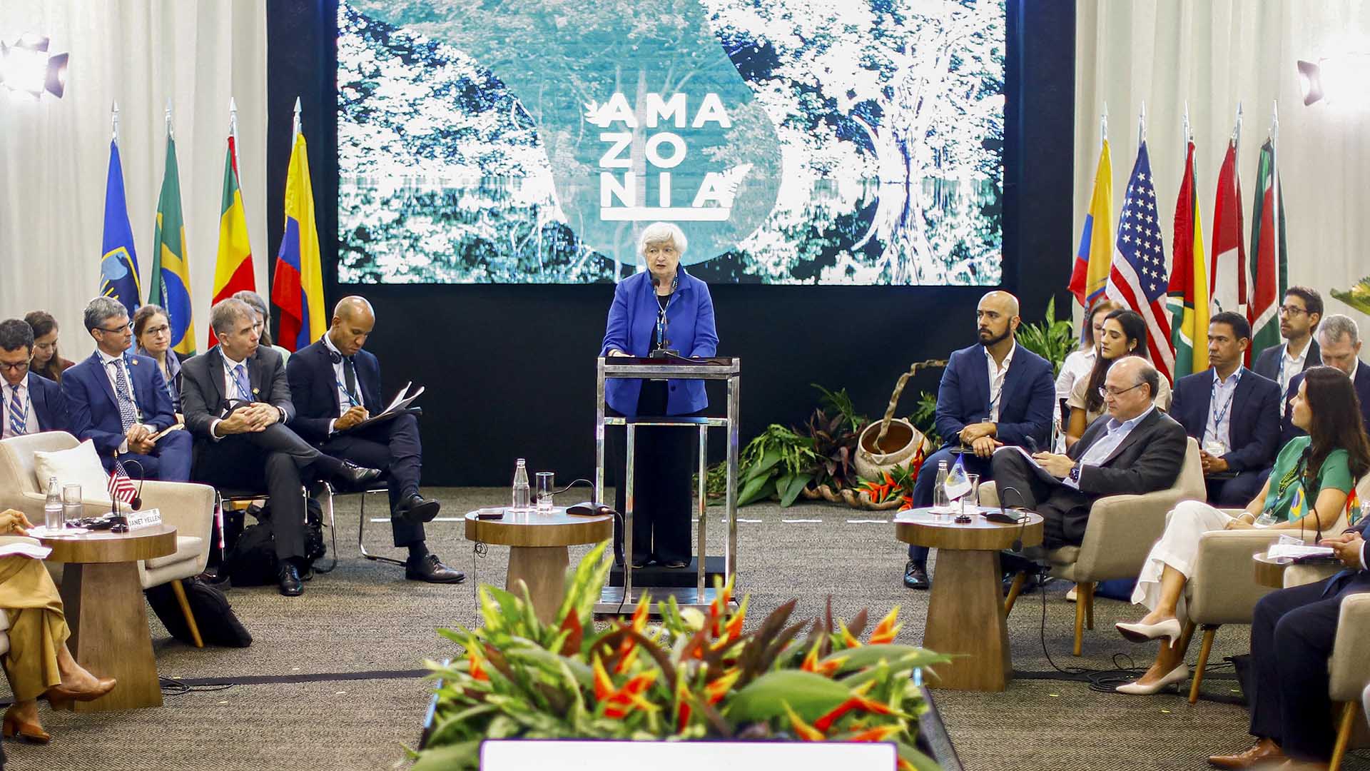 Janet Yellen stands behind a lectern in the center of a large room, with national flags from several South American countries and a screen reading Amazonia behind her.