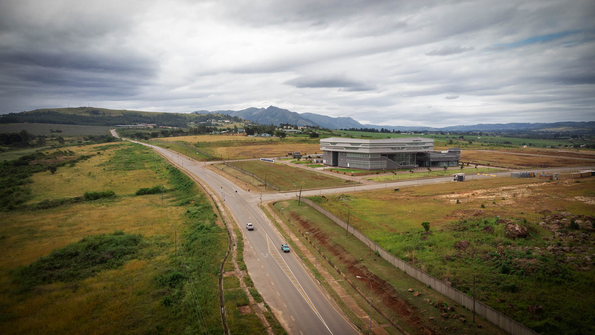 Wide photo showing a road passing through fields with a single, large building surrounded by empty land, with hills in the background.