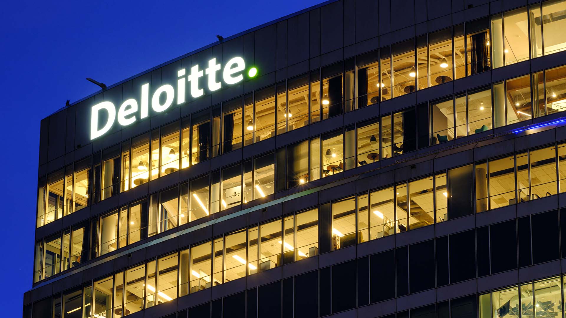 Top floors of an office building with windows illuminated against a night sky and a lit-up sign reading Deloitte