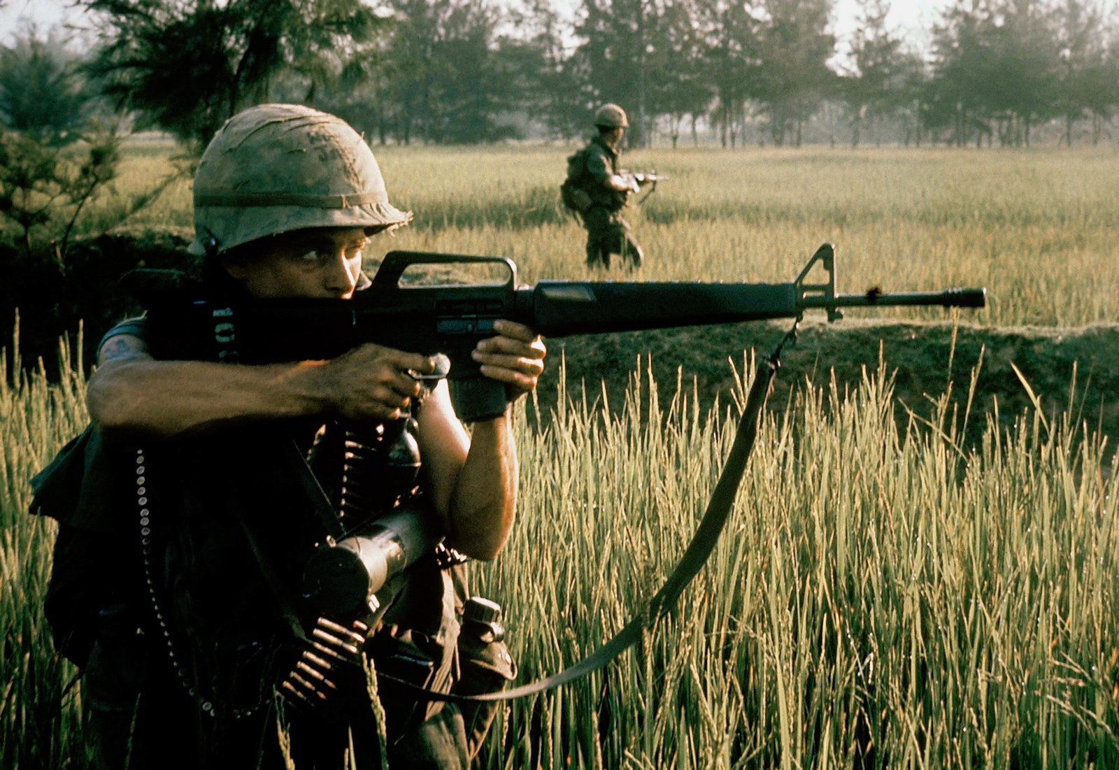 A soldier fires a rifle in the grassy fields of My Lai with one of his peers scouting behind him