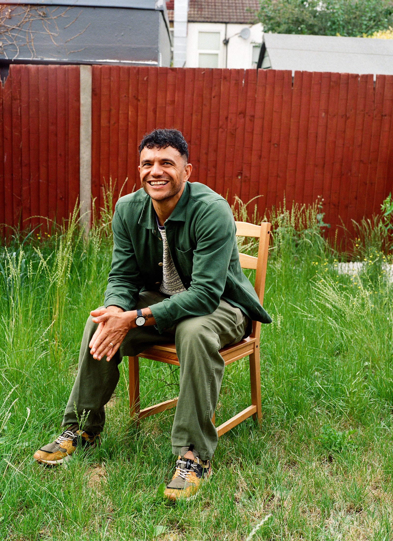 Raymond Antrobus sits on a chair in a yard wearing sneakers green pants and a green shirt. He has his hands together in...