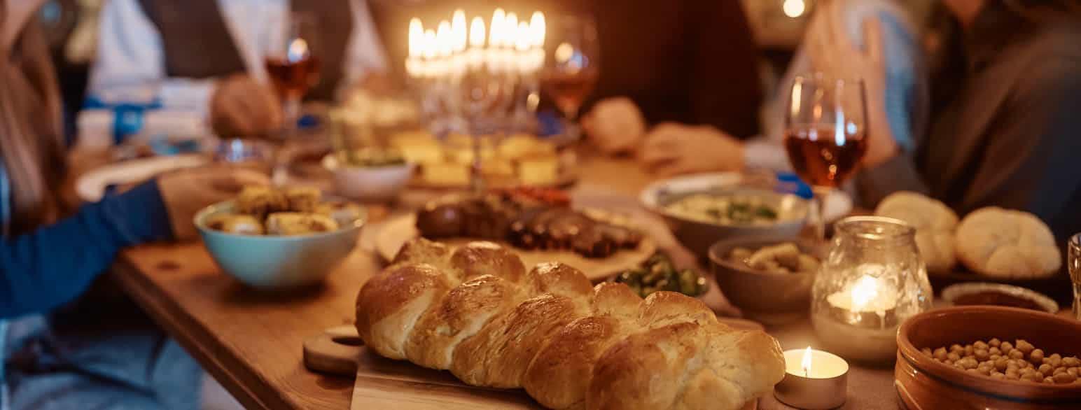 Et bord dekket med mat, tallerkner og glass. I forgrunnen en fletteloff. På bordet står også en lysestake med ni lys (en menorah). Rundt bordet sitter det folk. Foto