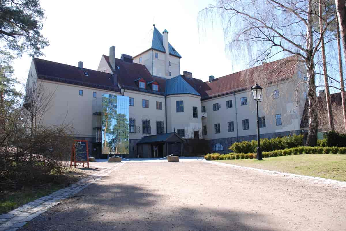 En tre-etasjes bygning med at tårn med to etasjer til midt på. En bred grusvei leder opp mot huset. Det er en plen med busker og en bjørk på plenen. Foto