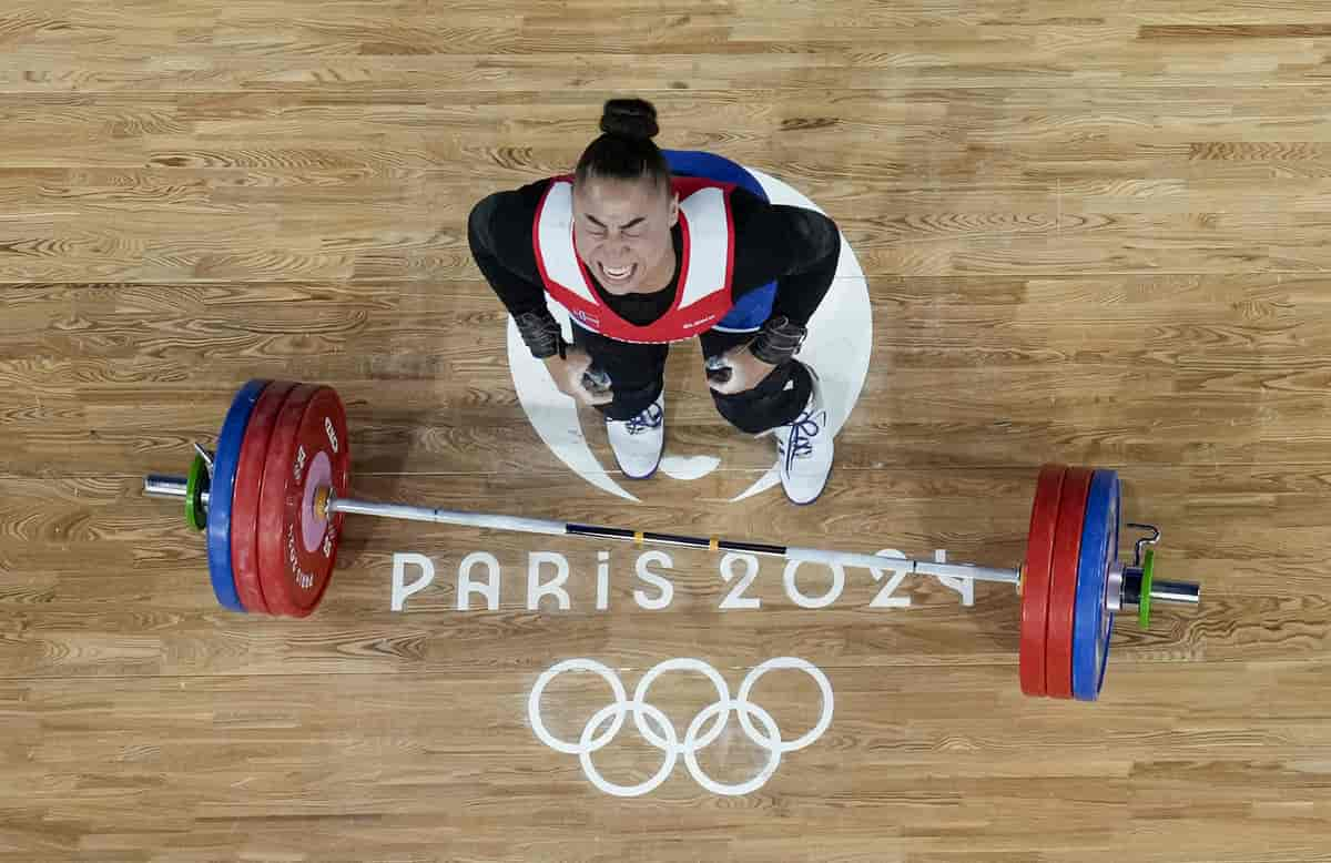 Foto av Koanda tatt ovenfra. Hun står på en parkett med logoen til OL i Paris og teksten «Paris 2024» på. Foran henne ligger en vektstang med vekter i hver ende. Hun ser litt oppover mot kamera og har lukka øyne og et stort glis.