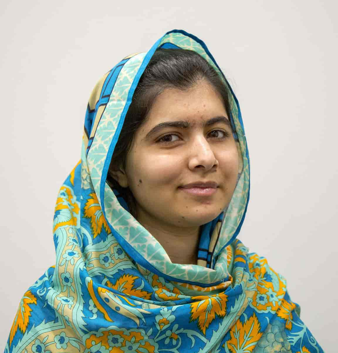 Malala Yousafzai er fotografert delvis fra siden, fra skuldrene og opp, bakgrunnen er hvit. Hodet og skuldrene er dekket av et sjal med blått og gult mønster. Hun smiler forsiktig med lukket munn. 