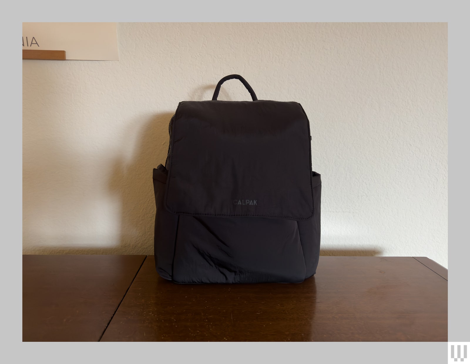 Smooth black backpack with small handle sitting on brown surface in front of offwhite wall