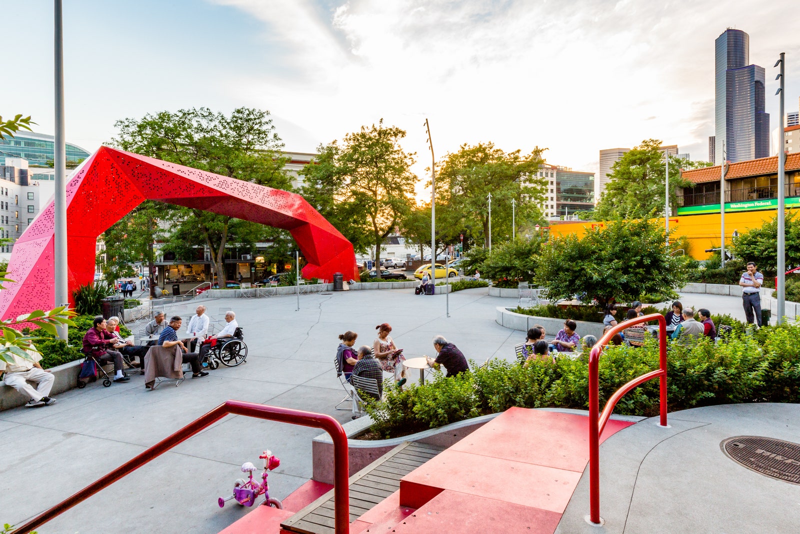 People sitting in a small park with a large red arch at the entrance and buildings in the background