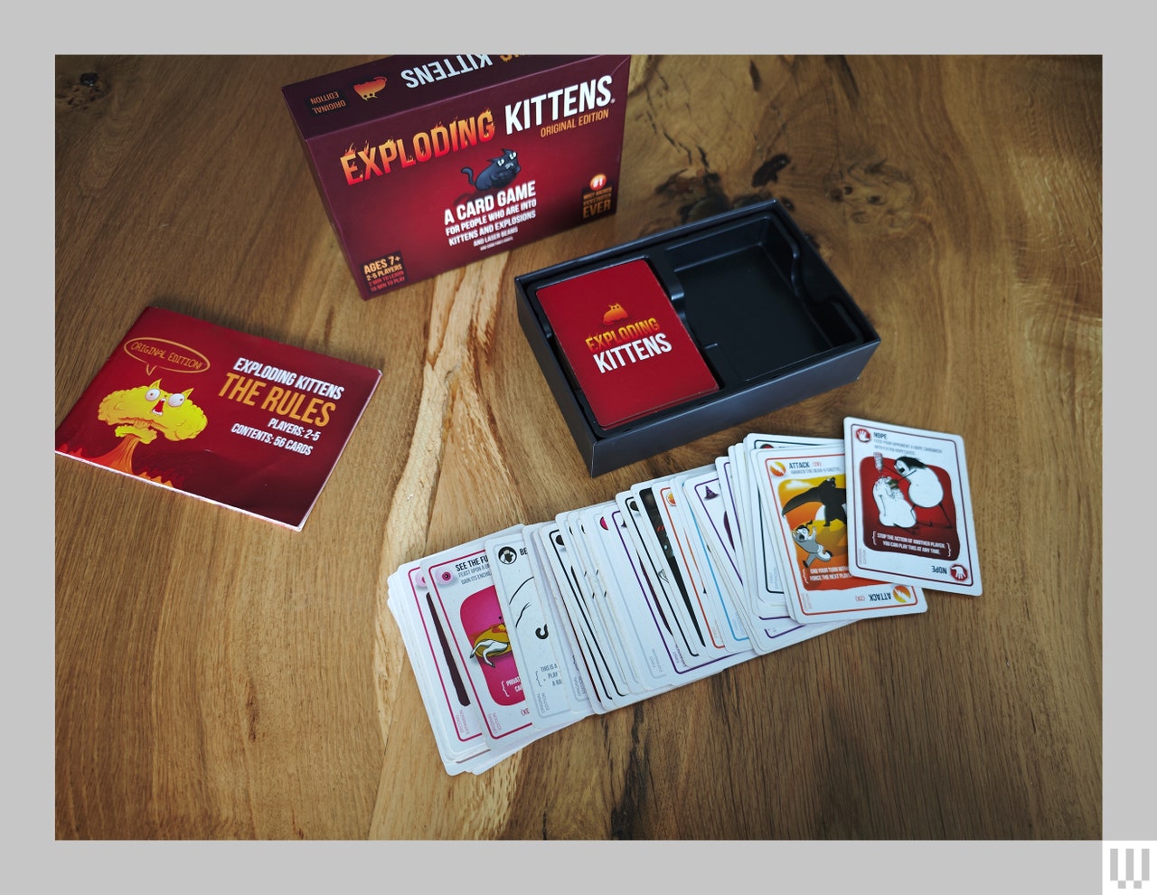 Contents of a card game spread out on a wooden surface. In the back is the red box and pamphlet, in the center are the cards spread out in a line on top of each other, and to the right is the empty box.