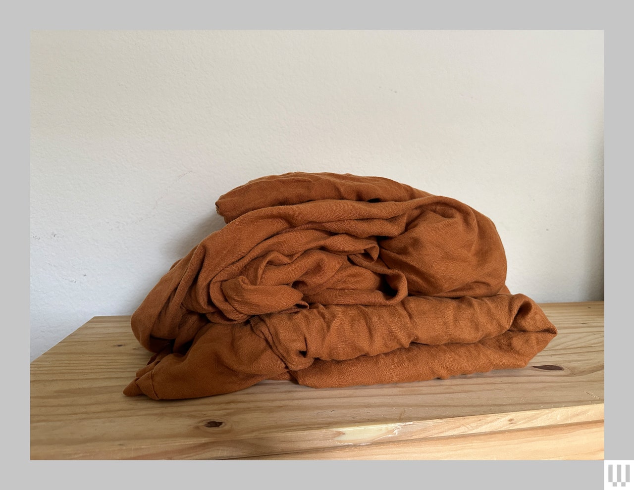 Balled up rust colored linen sheets sitting on a wooden surface in front of a white wall