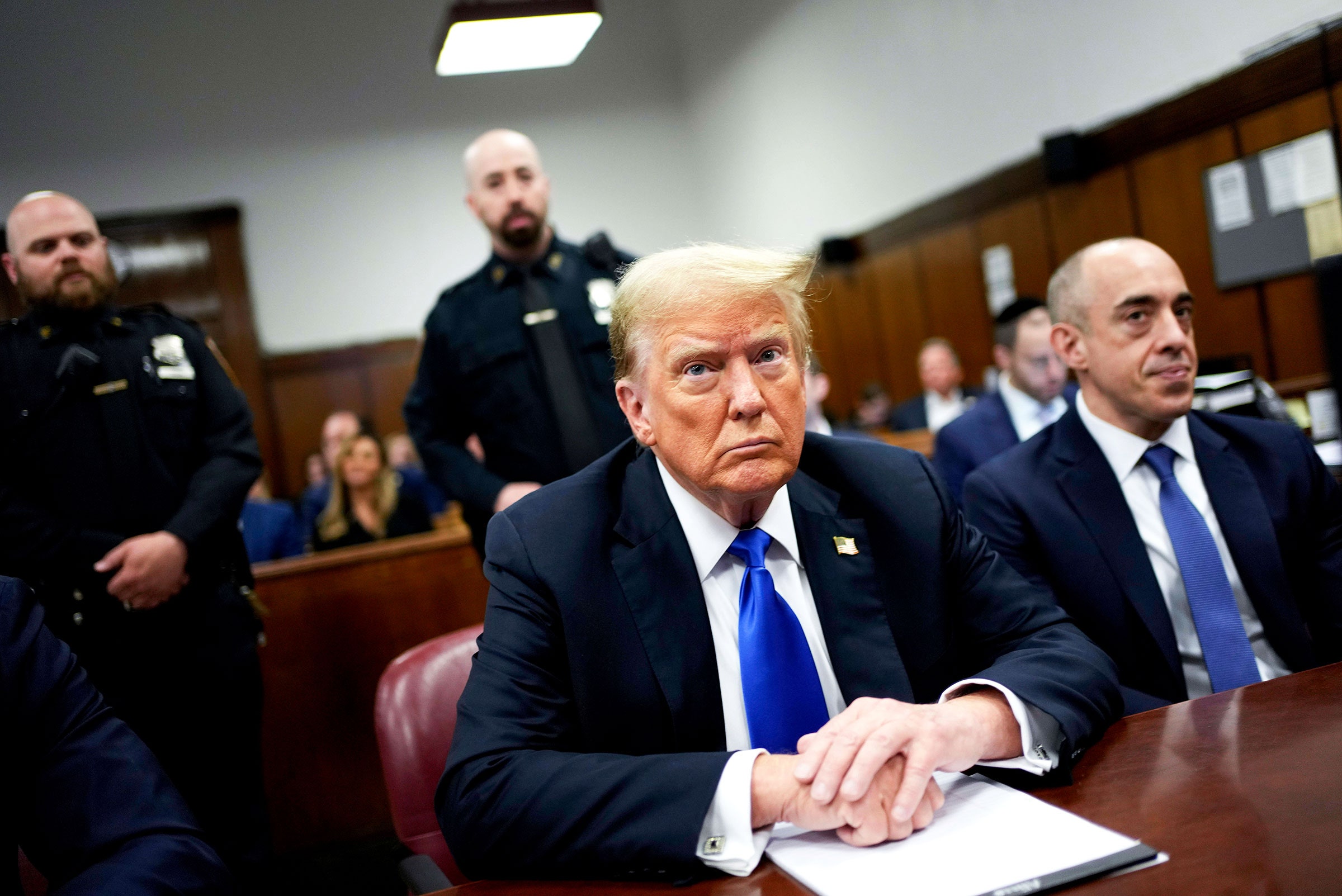 Donald Trump and his lawyer sitting at a desk in a court room with officers and onlookers behind them