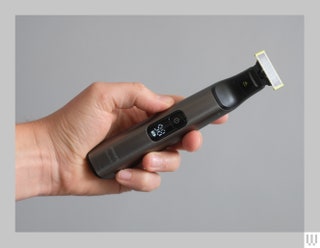 Hand holding a slim grey and black beard trimmer showing the front side with a small screen