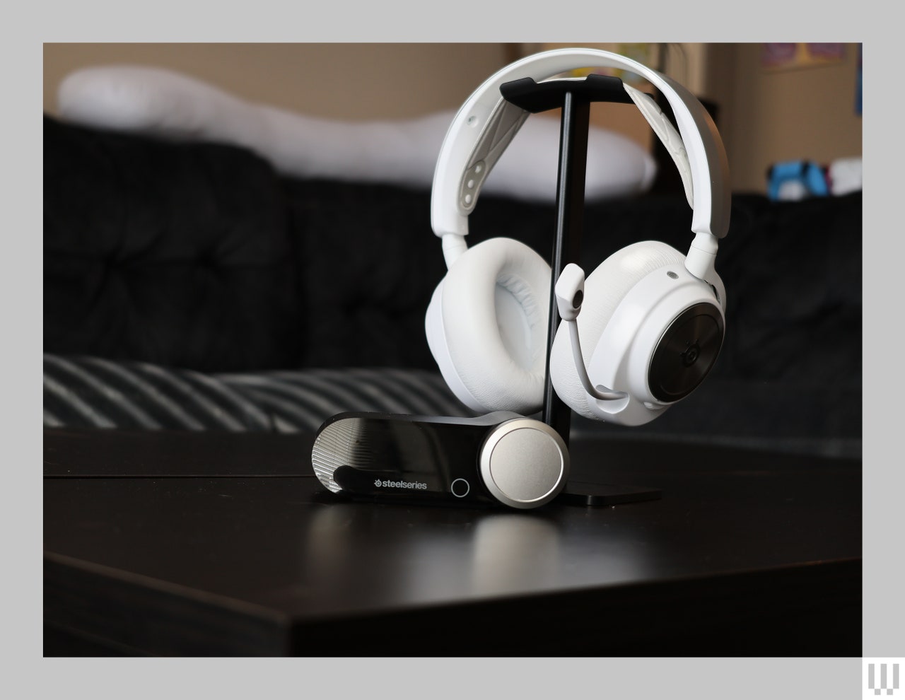 White headset with cushioned ear cups and extended mic hooked on a tabletop stand. Oval-shaped device to customize the audio is in front of the stand.