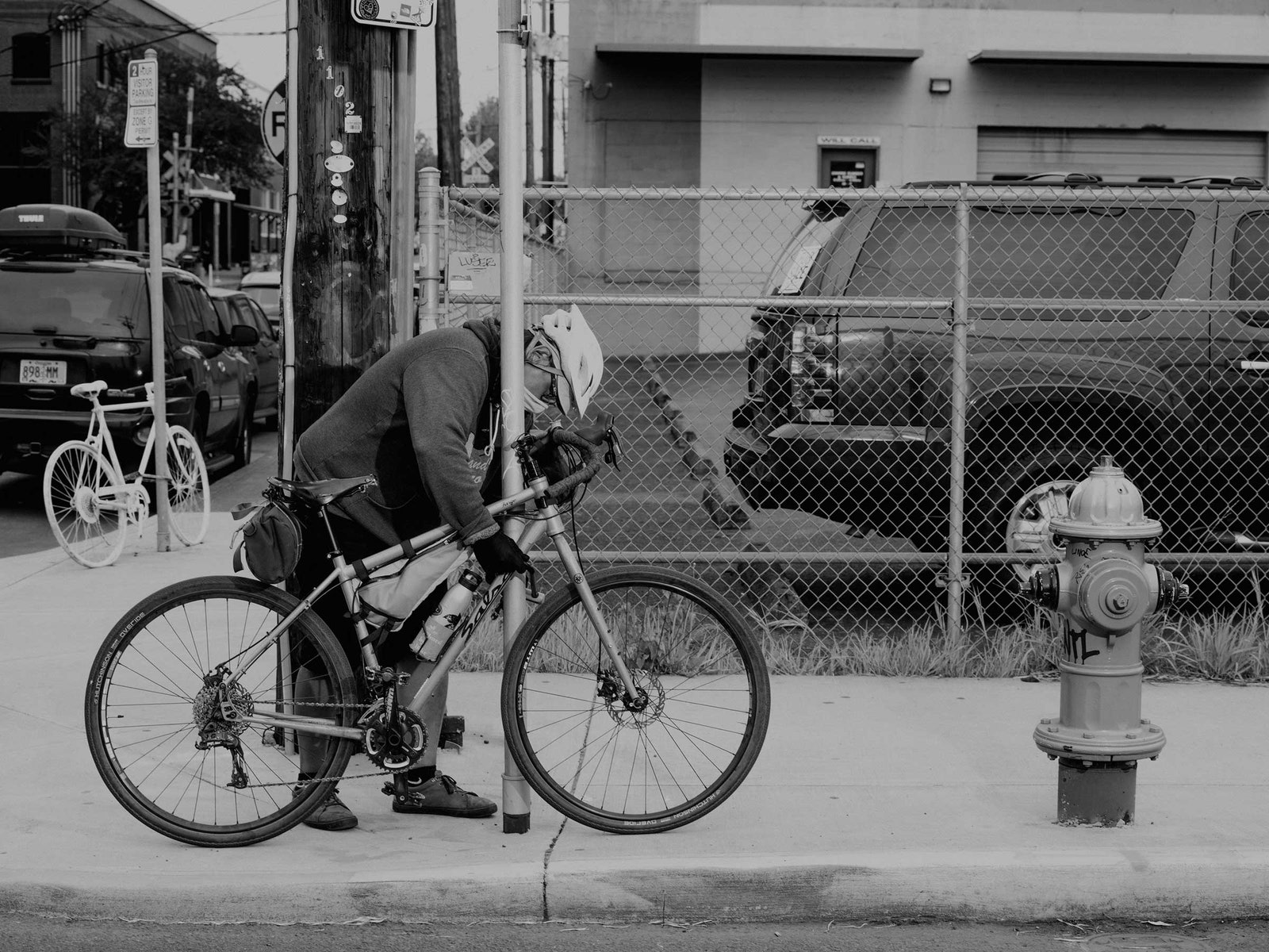 A black and white photograph of a person chaining a bicycle to a street sign with cars and buildings in the background