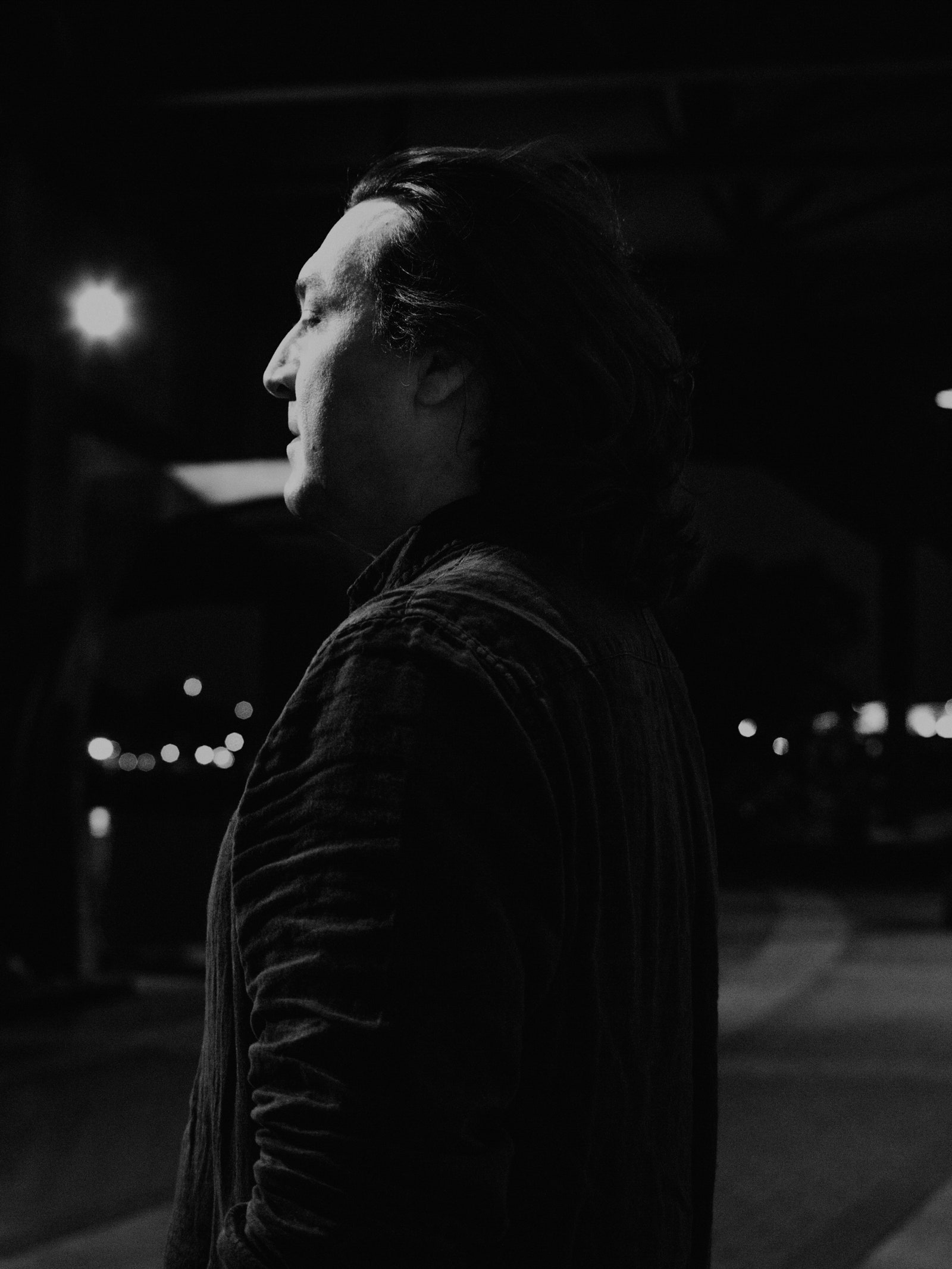 Black and white portrait of a person with long dark hair closing their eyes while standing on a street at night