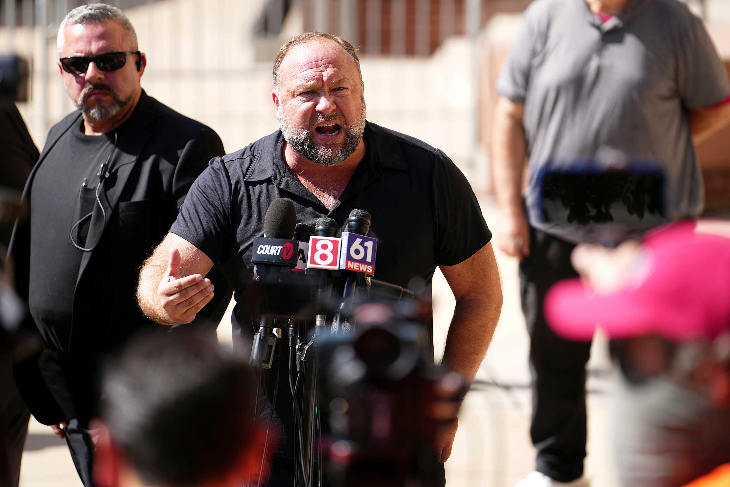 Alex Jones looking disgruntled while speaking into several microphones at a press conference outside
