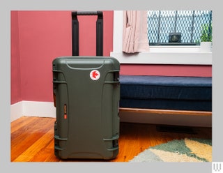 Hard shell luggage with thick handle protruding from the top with a bed and pink walls in the background