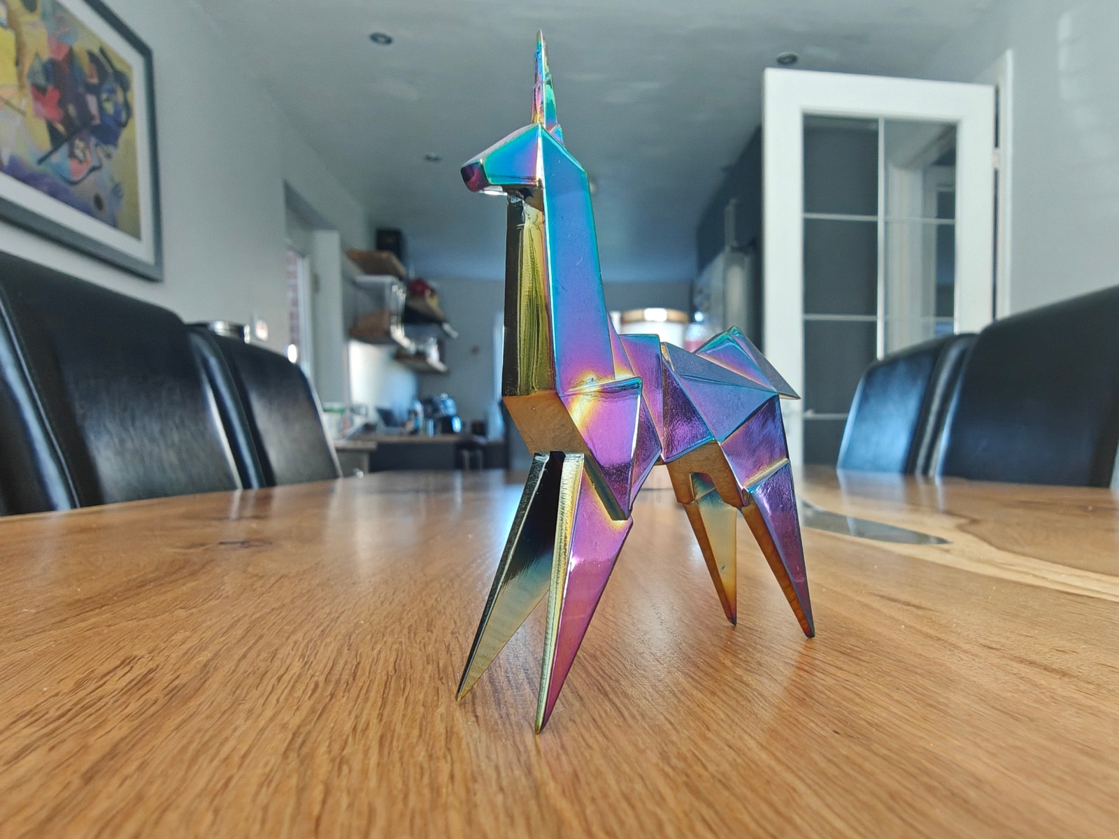 Chrome iridescent mini sculpture of an angular unicorn on a wooden table with black chairs on either side of the table